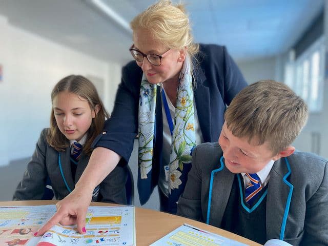 A teacher points out a section of a textbook on a class desk of two students.